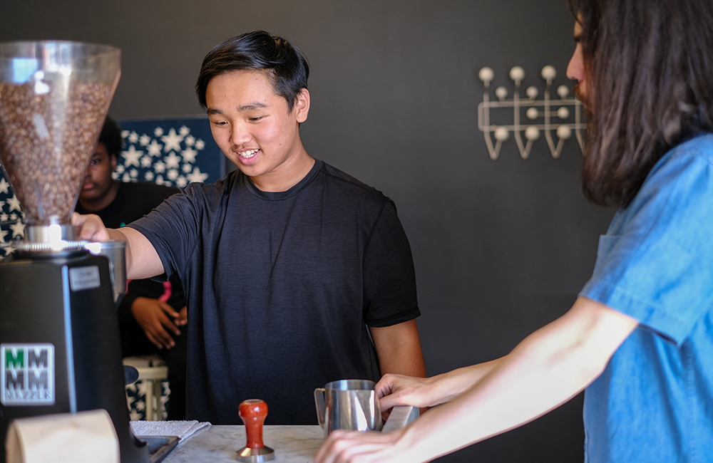 Teen boy operates coffee machine as barista looks on