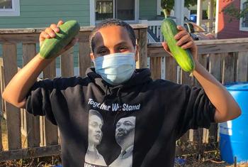 Young man in face covering holds up two cucumbers from NewHolly youth garden