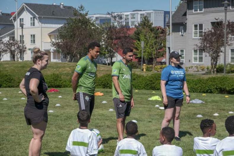 Two Seattle Sounders players talking to kids listening and seated on a field