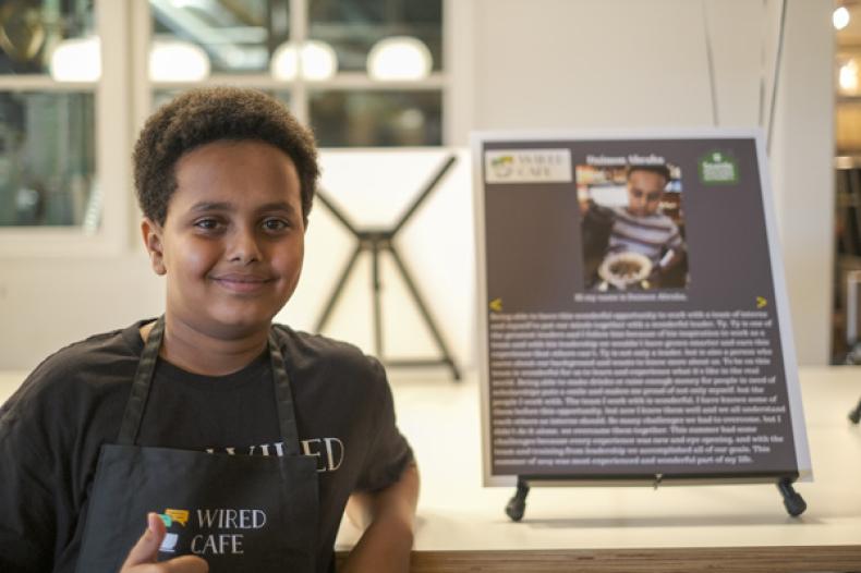 Teenage boy stands next to panel describing youth program to learn about operating a cafe