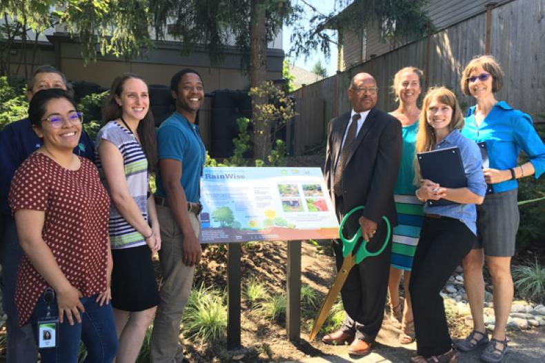Group of smiling people stand near panel describing Rainwise stormwater collection program outside apartment building