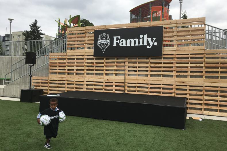 Child carrying two soccer balls in front of outdoor stage