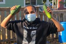 Young man in face covering holds up two cucumbers from NewHolly youth garden